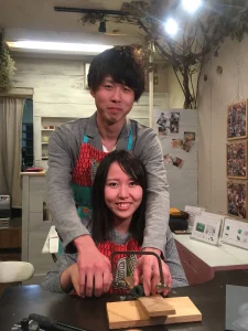 手作り結婚指輪の制作体験で、男性が女性の手に添えて糸鋸で地金を切る様子。工房で笑顔のカップルがふたり一緒に指輪づくりに取り組む温かな風景