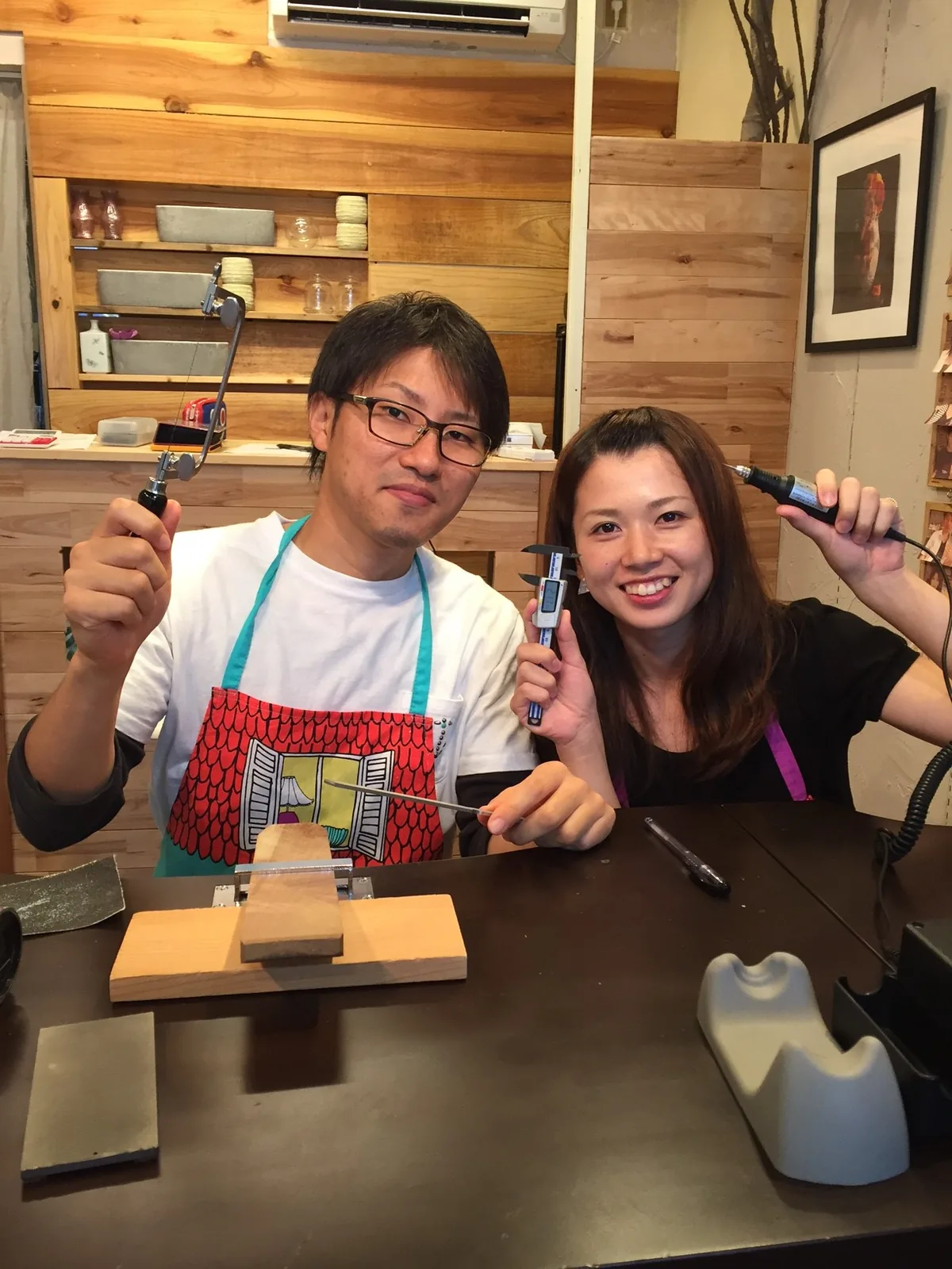 手作り結婚指輪の制作体験中、糸鋸やノギスなどの道具を手に笑顔を見せるカップル。ふたりで指輪をつくる工房のあたたかな雰囲気
