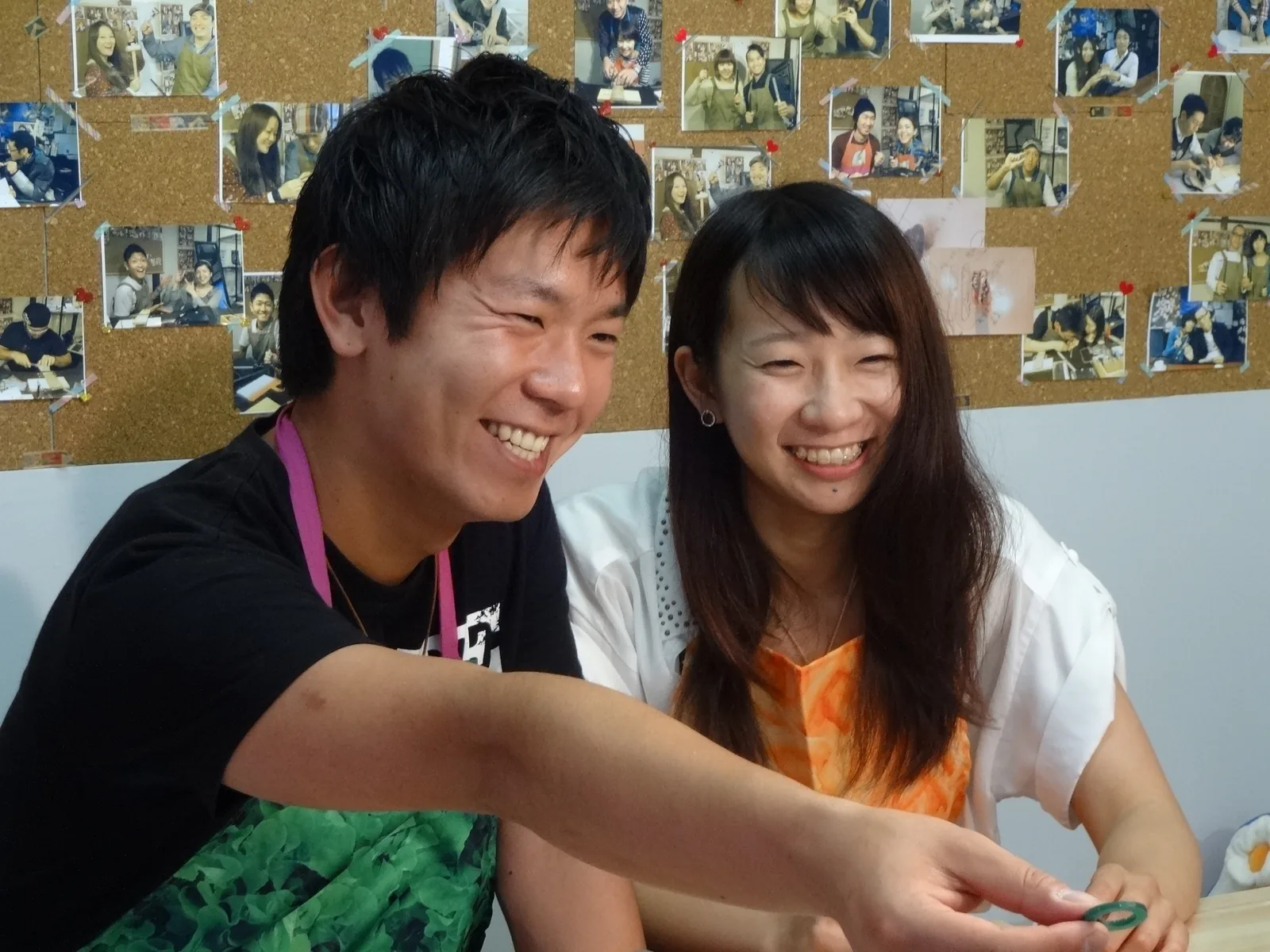 手作り結婚指輪の工房で、削り出したワックス原型を見つめて笑い合うカップル。お客様の写真が並ぶ壁を背に制作体験を楽しむ自然な表情